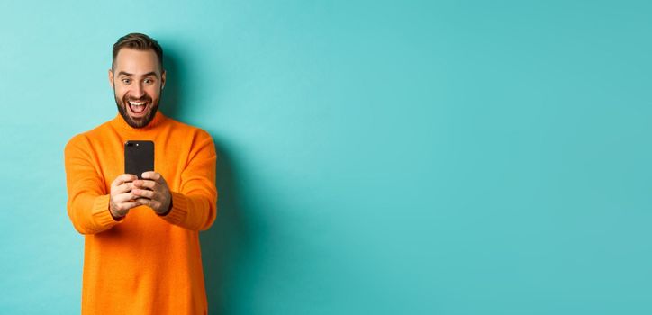 Image of man reading awesome message on mobile phone, looking at smartphone screen with amazement, standing over light blue background.