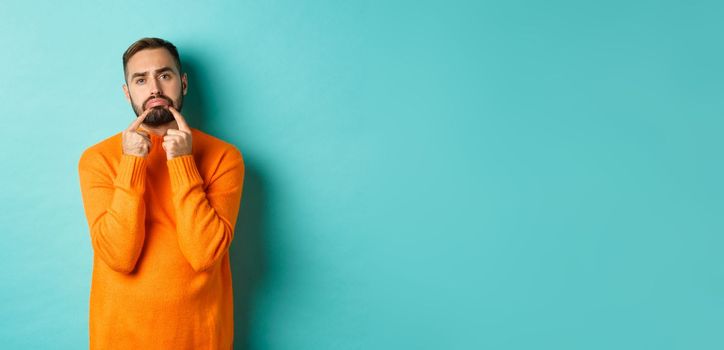 Image of gloomy bearded man, making sad face and frowning, standing upset in orange sweater against turquoise background.