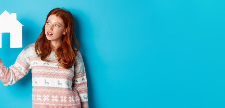 Real estate concept. Image of cute redhead girl looking curious at paper house model, thinking of buying property, standing in sweater against blue background.