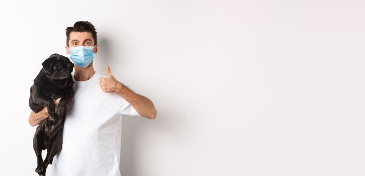 Covid-19, animals and quarantine concept. Young man in medical mask holding cute black pug dog, showing thumb up, like and approve, standing over white background.