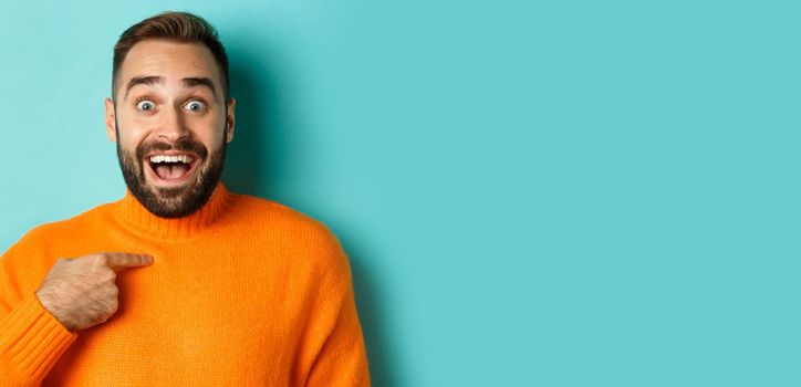 Close-up of happy young man pointing at himself with surprise and excitement, standing over light blue background.