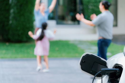 Focus closeup electric vehicle recharging battery from home electric charging station with blurred family in background. Renewable clean energy car for progressive eco awareness lifestyle concept.