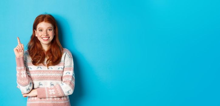 Excited redhead teen girl having an idea, raising finger and smiling, suggesting something, standing in sweater over blue background.