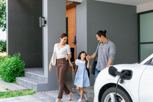 Progressive young parents and daughter with electric vehicle and home charging station. Green and clean energy from electric vehicles for healthy environment. Eco power from renewable source at home.