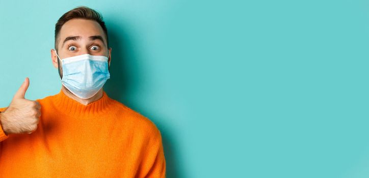 Covid-19, social distancing and quarantine concept. Close-up of cheerful man in medical mask showing thumb-up, light blue background.