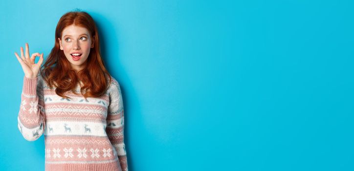 Silly redhead woman in sweater, showing okay sign and staring at upper left corner logo, standing over blue background satisfied.