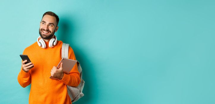 Handsome man student with headphones and backpack, holding digital tablet and smartphone, looking dreamy at upper left corner, standing against turquoise background.