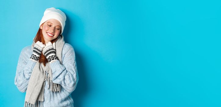 Winter and holidays concept. Cute redhead girl blushing and looking away dreamy, wearing warm beanie, sweater and scarf with gloves, standing over blue background.