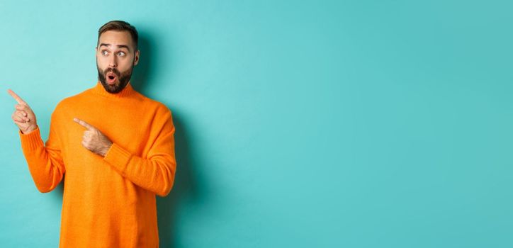 Surprised and curious guy looking at promo, pointing at upper left corner copy space, standing in orange sweater, turquoise background.