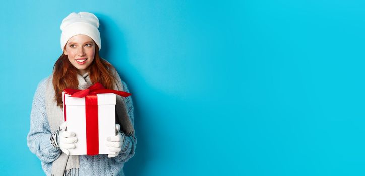 Winter holidays and Christmas sales concept. Thoughtful redhead woman standing in white beanie and gloves, holding holiday gift and looking left, thinking, standing over blue background.