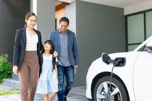 Progressive young parents and daughter with electric vehicle and home charging station. Green and clean energy from electric vehicles for healthy environment. Eco power from renewable source at home.