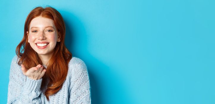 Lovely redhead female model smiling, sending air kiss at camera, standing against blue background.