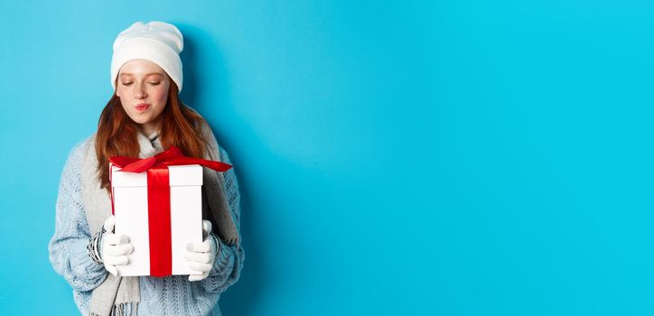 Winter holidays and Christmas sales concept. Intrigued redhead girl holding present, curiously staring at box with gift, trying guess what inside, standing over blue background.