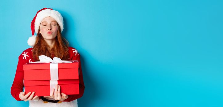Happy holidays and Christmas concept. Cute redhead girl holding presents and pucker lips for kiss, wearing santa hat and funny sweater, blue background.