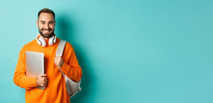 Happy man with backpack and headphones, holding laptop and smiling, going to work, standing over turquoise background.