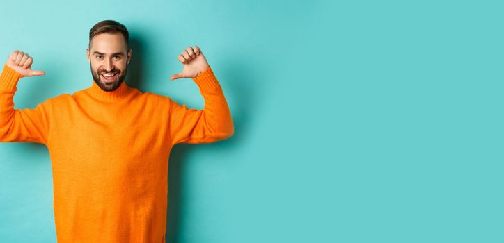 Handsome confident man pointing at himself, looking self-assured, standing in orange sweater against light blue background.