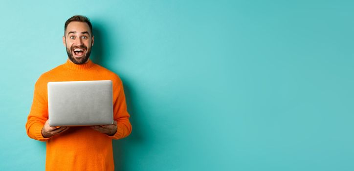 Happy man using laptop and looking excited at camera, standing with computer against light blue background.