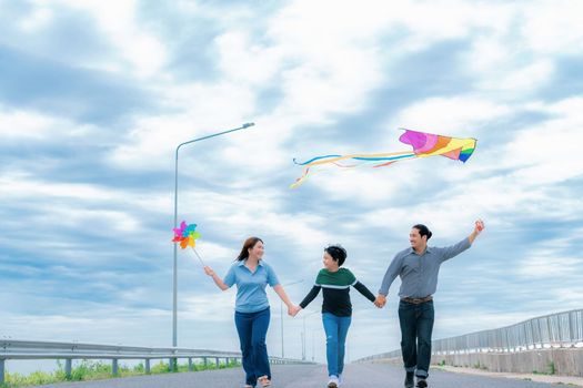 Progressive happy family vacation and carefree day concept. Young parents mother father and son run along and flying kite together road with enjoy natural scenic on scenery and clear sky background.