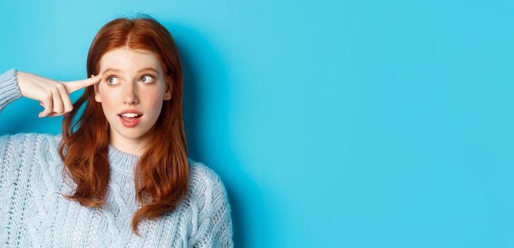 Close-up of cute redhead girl searching for solution, pointing at head and looking left at logo, standing over blue background.