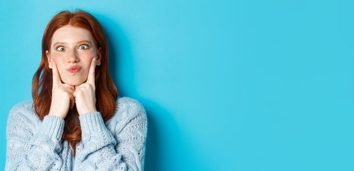 Close-up of funny redhead teen girl making faces, squinting and pocking cheeks, standing against blue background.