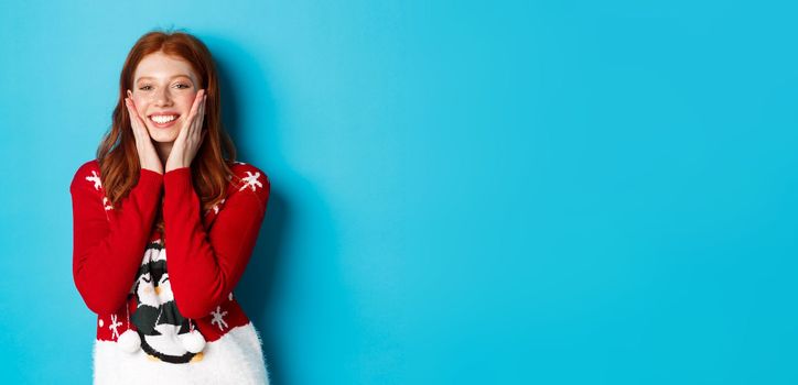 Winter holidays and Christmas Eve concept. Cheerful and cute redhead girl in xmas sweater, blushing and touching cheeks, smiling and rejoicing over blue background.