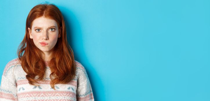 Close-up of serious-looking girl thinking, staring at camera troubled and grimacing, making hard choice, standing over blue background.