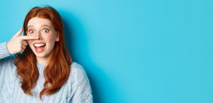 Close-up of silly redhead girl showing piggy nose, making funny grimaces and smiling, standing against blue background.