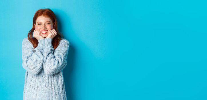 Lovely redhead teenage girl looking daydreaming, staring with admirationa sympathy at camera, standing against blue background.