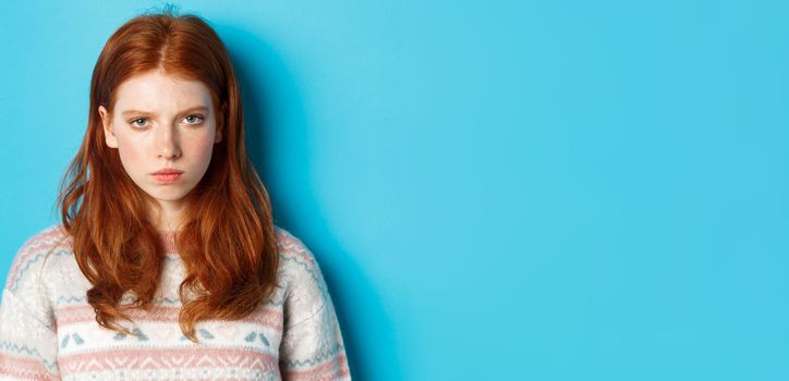 Close-up of angry redhead teenage girl looking pissed-off at camera, frowning and sulking mad, standing over blue background.