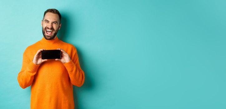 Photo of happy adult man laughing and showing funny thing on mobile phone screen, standing in orange sweater over turquoise background.