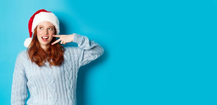 Winter holidays and Christmas Eve concept. Happy teenage girl with red hair, wearing santa hat, enjoying New Year, showing peace sign and gazing left at promo, standing over blue background.