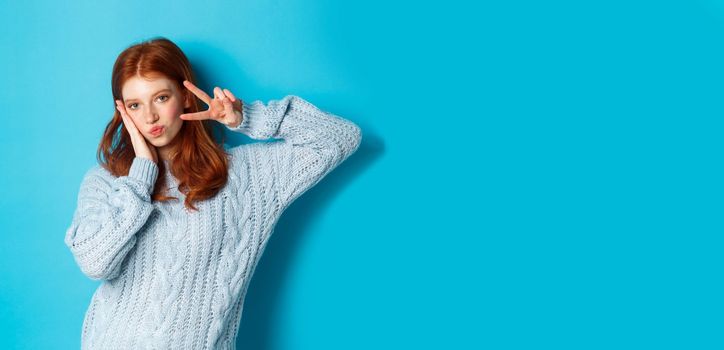 Modern teen girl with red hair, showing peace sign and posing in sweater against blue background.