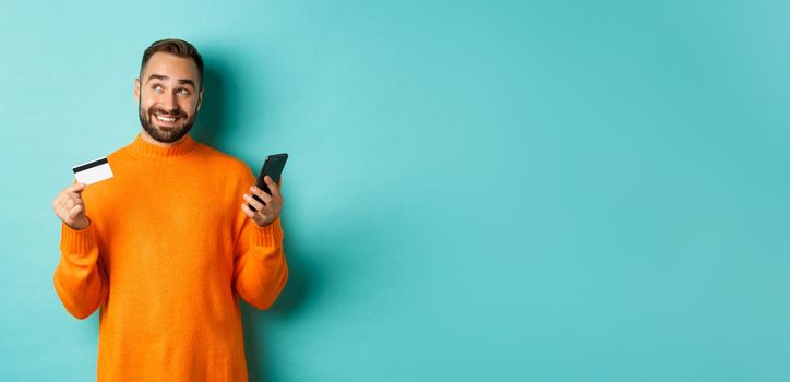 Online shopping. Handsome man thinking, holding smartphone with credit card, paying in internet store, standing over light blue background.