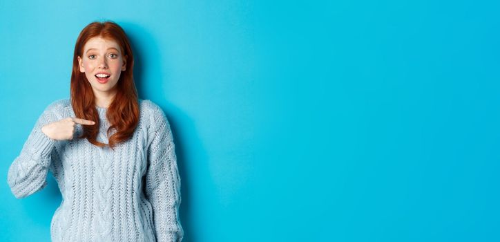 Hopeful redhead girl pointing at herself, standing over blue background.