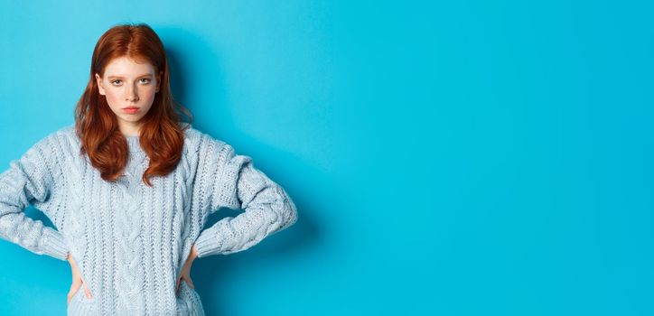 Redhead girl staring angry and displeased at camera, frowning upset, standing against blue background.