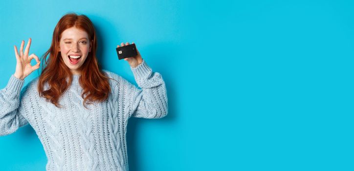 Happy redhead girl in sweater showing credit card and okay sign, recommending bank offer, standing over blue background.