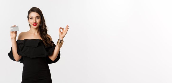 Beauty and shopping concept. Gorgeous woman in luxury jewelry and black dress, showing okay sign and credit card, standing over white background.