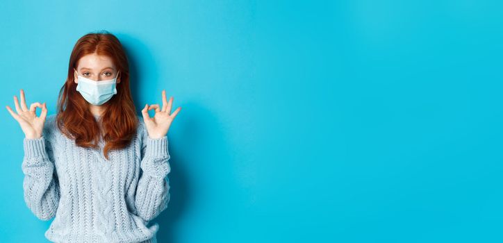 Winter, covid-19 and social distancing concept. Satisfied young redhead woman in face mask showing alright, okay gestures and looking pleased, standing blue background.