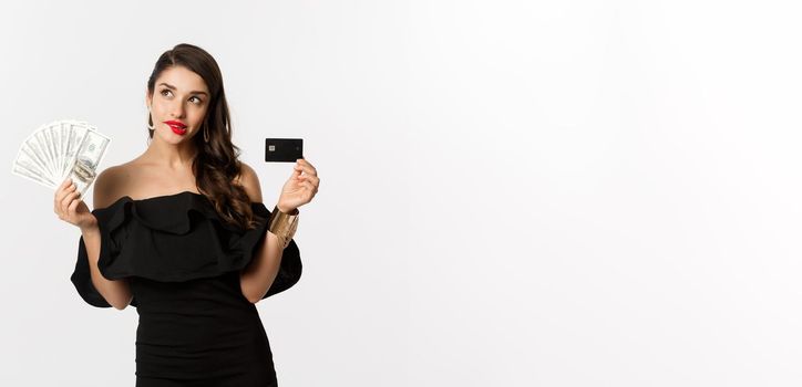 Fashion and shopping concept. Elegant woman in black dress, red lipstick, holding money and credit card, making choice, thinking, standing over white background.