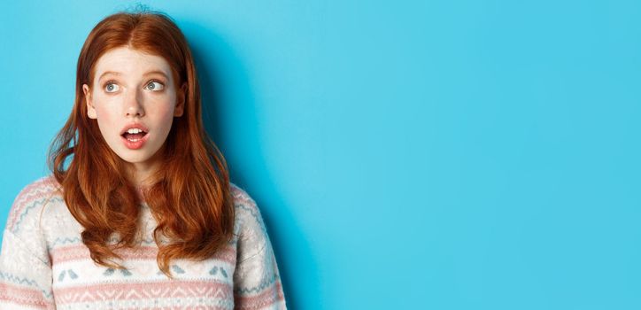 Close-up of impressed redhead girl staring left amazed, open mouth in awe and glancing at promo, standing in winter sweater against blue background.