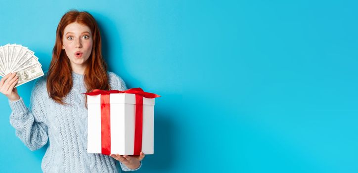 Christmas and shopping concept. Excited redhead girl looking at camera, holding big New Year gift and dollars, buying presents, standing over blue background.