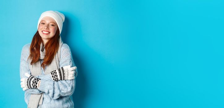 Winter and holidays concept. Smiling redhead girl in beanie, gloves and sweater getting warm after going outside, standing over blue background.