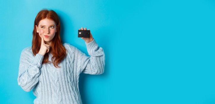 Image of thoughtful redhead girl thinking about shopping, showing credit card and pondering, standing over blue background.
