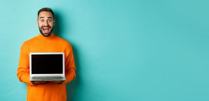 Handsome bearded man in orange sweater showing laptop screen, demonstrating promo, standing over light blue background.