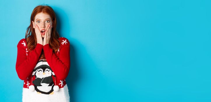 Winter holidays and Christmas Eve concept. Impressed and speechless redhead girl gasping, staring at camera with disbelief, standing in xmas sweater against blue background.