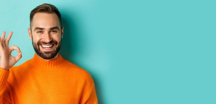 Close-up of assertive young man assuring everything ok, showing okay sign and smiling, yes or positive answer, standing over light blue background.