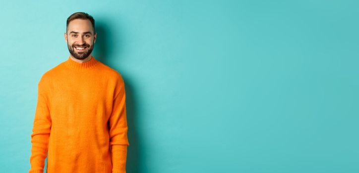 Lifestyle. Handsome man in orange sweater smiling at camera, happy face, guy cheerful expression, standing over turquoise background.