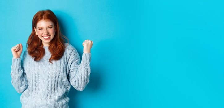 Satisfied redhead girl achieve goal and celebrating, making fist pump gesture and smiling delighted, triumphing of win, standing against blue background.