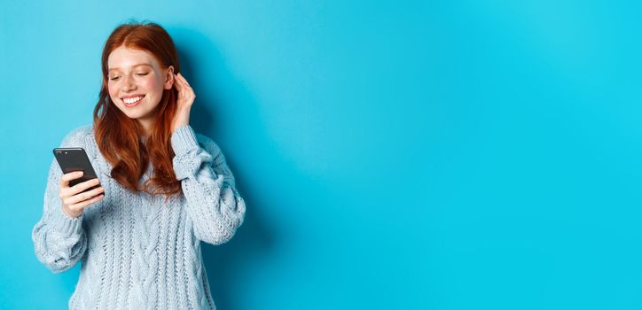 Technology. Happy redhead girl looking pleased at mobile phone, reading compliment in message, smiling and tuck hair behind ear, standing in sweater over blue background.