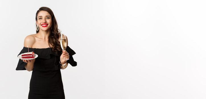 Celebration and party concept. Happy woman holding birthday cake and drinking champagne, smiling while standing in black dress against white background.